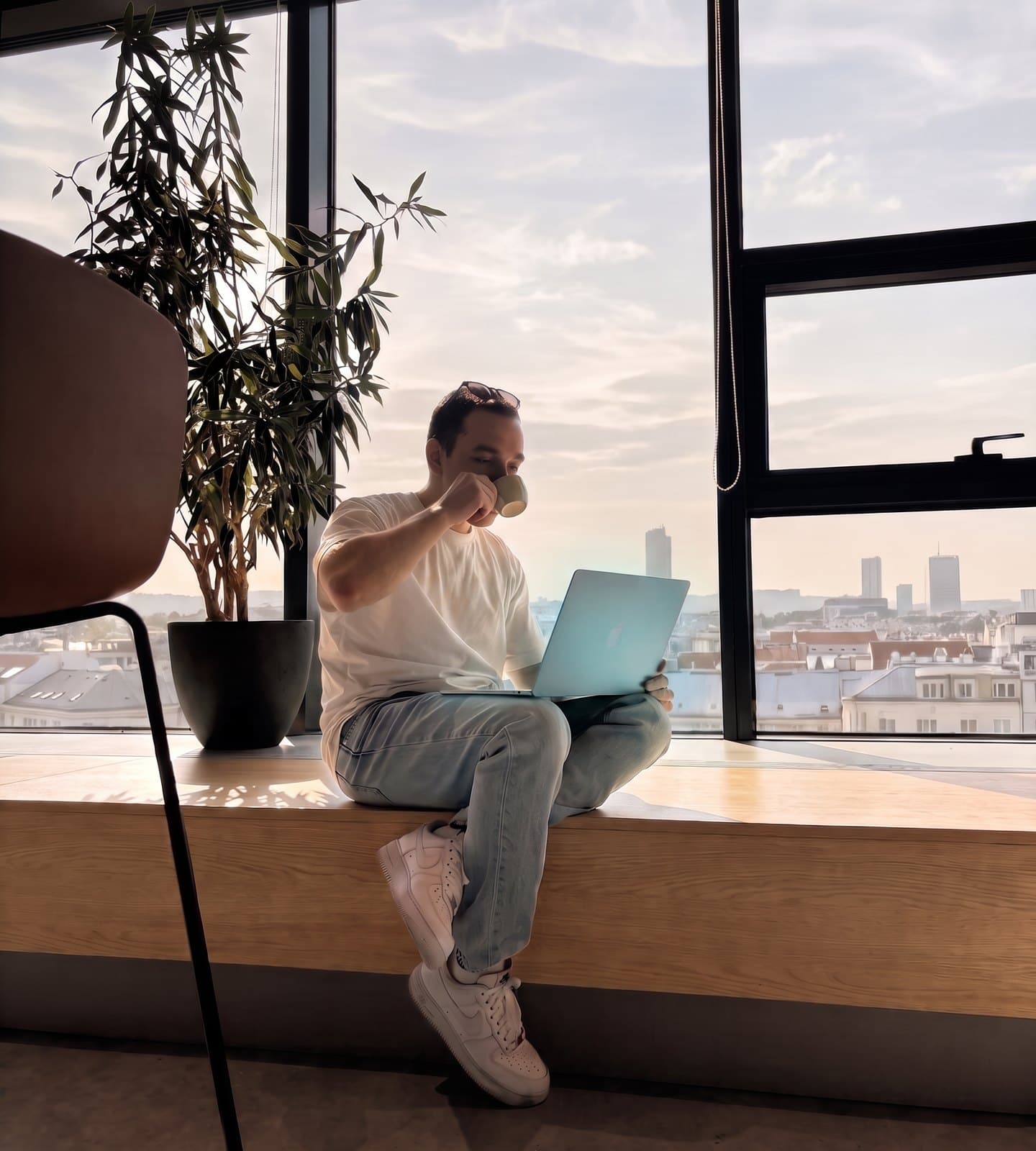 An engineer sitting cross-legged on a window bench with a laptop and a cup of coffee, city rooftops behind him